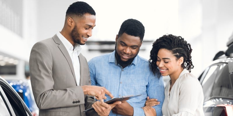 A car sales setting where three people stand beside a vehicle