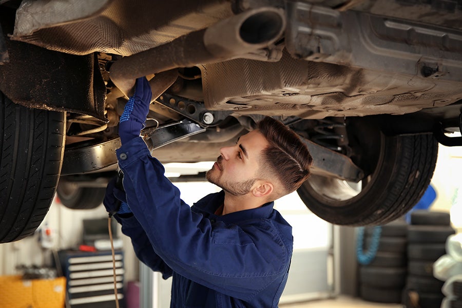 A mechanic in a blue uniform examines the underside of a raised car in a garage