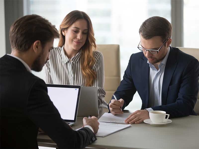 Two people review and sign documents at a table while another person sits