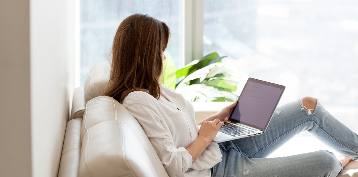 A person sitting on a light-colored sofa uses a laptop in a bright room with large windows